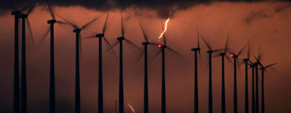The silhouette of spinning wind turbines is backlit by a bolt of lightning in a thunderstorm at night. Quillete | James Alexander Michie